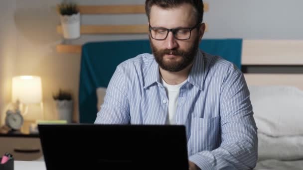 男は自宅のコンピュータで働いている。ひげを生やした男は、ラップトップキーボードのベッドルームとタイプのテーブルに座っています。自宅で働く、フリーランスのコンセプト — ストック動画