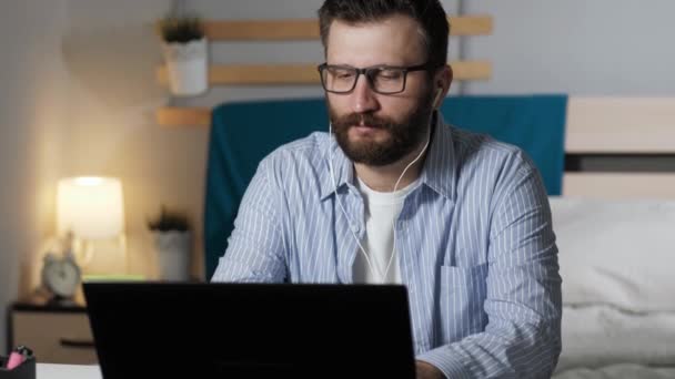 O homem ouve música e trabalha no computador em casa. O cara barbudo nos fones de ouvido se senta à mesa no quarto ouvindo música e digitando no teclado do laptop. Trabalho em casa, conceito freelance — Vídeo de Stock