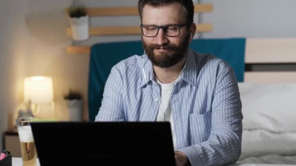 El hombre bebe cerveza escucha música y trabaja en casa. El barbudo con auriculares se sienta a la mesa en el dormitorio, trabaja en el portátil, bebe cerveza y escucha música. Trabajo en casa, concepto freelance — Vídeo de stock