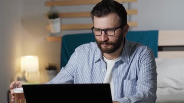 O homem trabalha no computador e bebe cerveja. O tipo barbudo senta-se à secretária no quarto a escrever no teclado do portátil e a beber cerveja. Trabalho em casa, conceito freelance — Vídeo de Stock