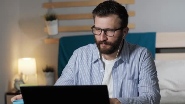 El hombre que trabaja hablando en la conexión de vídeo y bebe café. Tipo barbudo se sienta en el escritorio en el dormitorio hablando en conexión de vídeo y beber café o té. Trabajo en casa, concepto freelance — Vídeo de stock