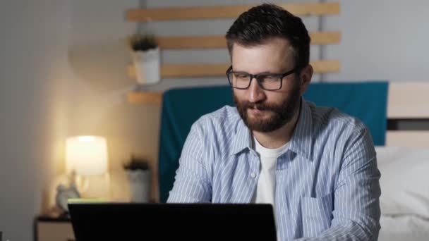 L'homme se réjouit à la fin du projet. Barbu gars assis au bureau dans la chambre à coucher tapant sur le clavier portable casse et applaudit joyeusement ses mains et lève les mains vers le haut. Travail à domicile, concept freelance — Video