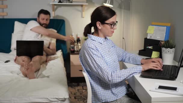 夫と妻は自宅でオンラインで働いています 女性は机に座って ノートパソコンのキーボードに何かを入力し 男はベッドルームでベッドの上に横たわって ビールを飲み ノートパソコンを使用します クローズアップとスローモーション — ストック動画
