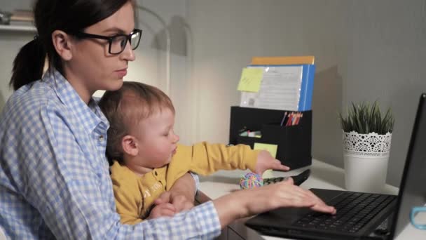 La chica trabaja en la computadora. Mujer atractiva se sienta en el escritorio escribiendo en el teclado del ordenador portátil y sostiene en sus brazos a un niño pequeño que interfiere con el trabajo — Vídeo de stock