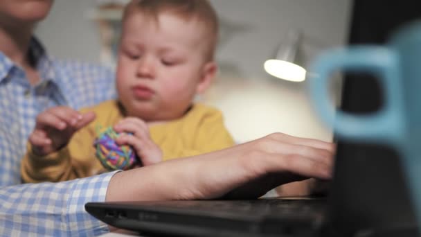 La mano femenina está escribiendo en la computadora portátil, en el fondo, el niño pequeño se sienta en los brazos de su madre y juega con artículos de papelería. De cerca. — Vídeos de Stock