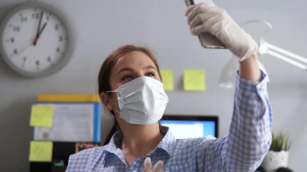 Chica con máscara y guantes se toma selfie. Mujer joven en guantes de látex blanco y máscara quirúrgica en su cara está en la oficina o en el lugar de trabajo en el apartamento y toma selfie en el teléfono — Vídeo de stock