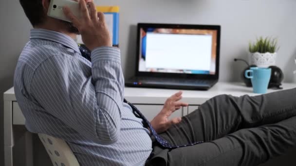 Υπάλληλος γραφείου μιλάει στο τηλέφωνο. Γενειοφόρος άνδρας με πουκάμισο και γραβάτα κάθεται χαλαρός στο χώρο εργασίας στο γραφείο ή το διαμέρισμα, πόδια διπλωμένα στο τραπέζι, μιλώντας στο τηλέφωνο και πληκτρολογώντας κάτι σε φορητό υπολογιστή — Αρχείο Βίντεο