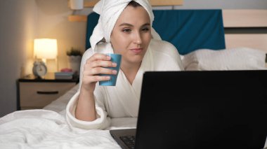 Kahve ya da çay içen ve dizüstü bilgisayarla çalışan bir kız. Yatak odasındaki beyaz bornozlu çekici kadın yatağa uzanıyor sıcak kahve ya da çay içiyor ve dizüstü bilgisayar kullanıyor. Yakın çekim görünümü