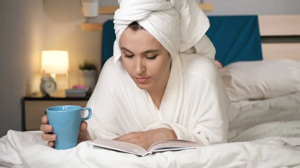女の子はコーヒーや紅茶を飲み 本を読む 彼女の頭の上にタオルが付いている白いバスローブの寝室の魅力的な女性は胃の飲み物熱いコーヒーか紅茶にあり 本を読み ページをひっくり返す クローズアップビュー — ストック写真