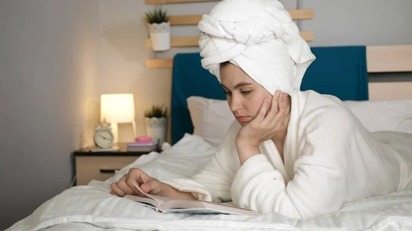 Girl lies on bed and reads book. Woman in bedroom with towel on her head and in white coat lies on her stomach in bed and turns over page of book. Medium shot view