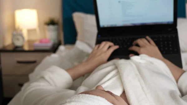 Girl lies on bed and types on laptop. Blurred view of woman in bedroom in white bathrobe lies on her back and types on laptop keyboard. Close-up view
