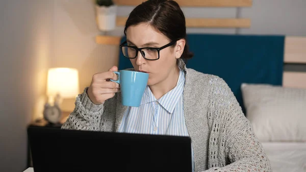 Girl drinks tea or coffee and works at computer. Woman with glasses alone in bedroom sits at desk typing on laptop keyboard and drinking tea or coffee. Work at home, freelance concept. Medium shot