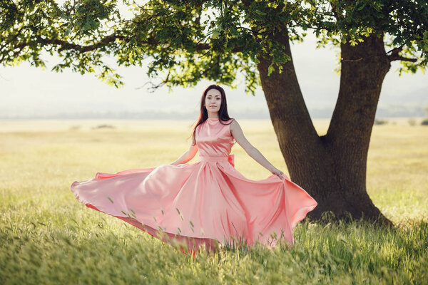 Beautiful woman posing in long pink dress on wheat field under big green tree