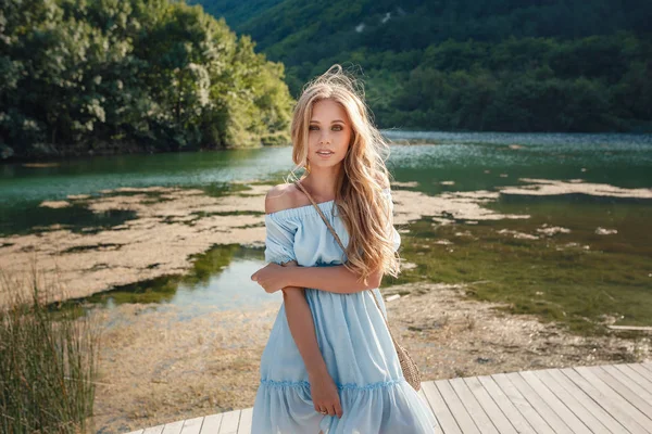 Young woman in blue dress standing alone on footbridge and staring at lake. Mist over water. Chilly morning. — Stock Photo, Image
