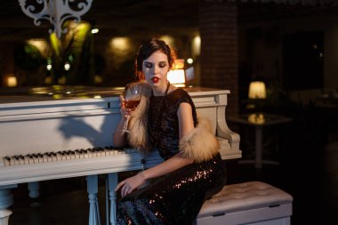 Portrait of 20s style festive beauty in a restaurant. Young beautiful woman in art-deco style, in black dress in a luxurious interere Chicago 20s, the era of gangsters. Singer at the white piano clipart