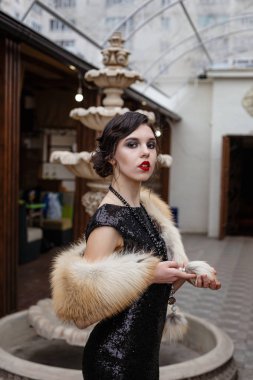 Bir restoranda 20 'li yılların tarzı şenlikli bir güzelliğin portresi. Sanat deco tarzı giyinmiş, siyah elbiseli, lüks bir interere sahip 20 'li yaşlarda, gangsterlerin çağı, çeşmede bir bardak ile.