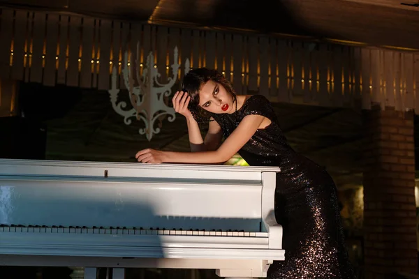 Portrait of 20s style festive beauty in a restaurant. Young beautiful woman in art-deco style, in black dress in a luxurious interere Chicago 20s, the era of gangsters. Singer at the white piano