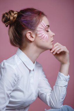 Bir çiçekle kameraya bakan güzel bir genç kadının portresi. Pembe arka planda çiçek Gerbera 'sı olan güzel bir kız yüzü. Cilt bakımı ve güzellik anlayışı. Beyaz bluz ve kot pantolon giyen şık ve şık küçük kız.