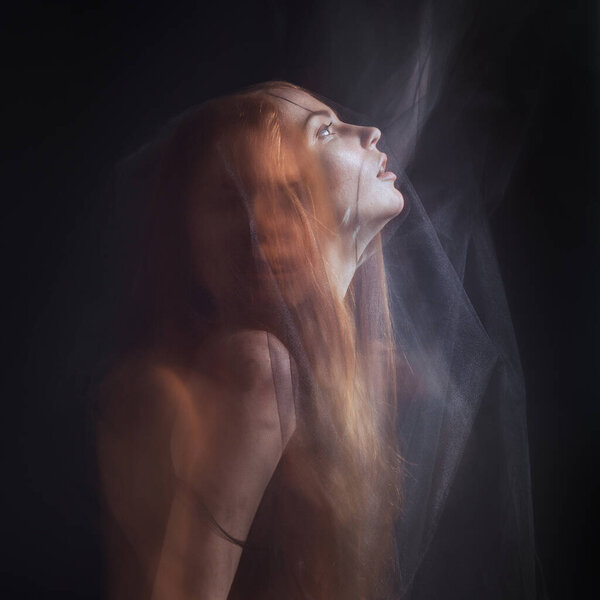 Beautiful model with red hair posing behind a black fabric and a veil in a studio. Technique mixed light