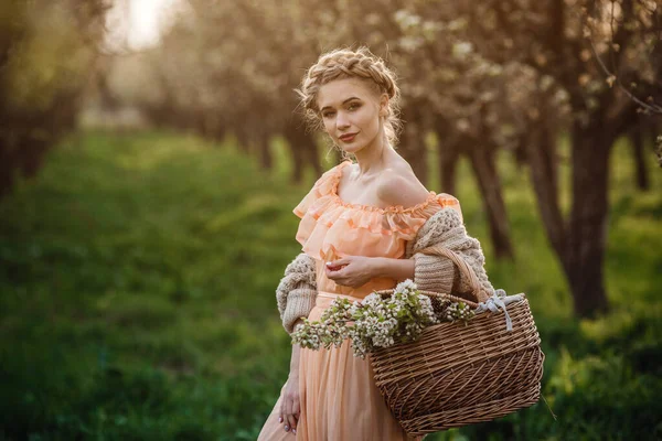 一个金发碧眼的女孩 穿着浅色的衣服 躺在花园子里 身着漂亮连衣裙和针织毛衣的女孩 带着一篮花 在梨花盛开的花园里欣赏夕阳 — 图库照片
