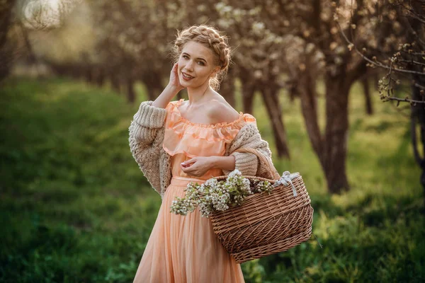 開花の庭の薄いドレスを着た金髪の女の子 美しいドレスとニットのセーターの女の子は 花のバスケットと梨の花の庭で夕日を楽しんでいます — ストック写真