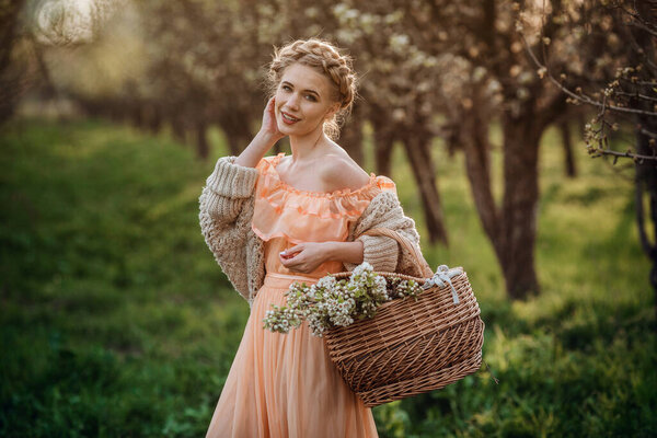 girl with blonde hair in a light dress in flowering garden. girl in a beautiful dress and knitted sweater enjoys the sunset in a pear flowering garden, with a basket of flowers