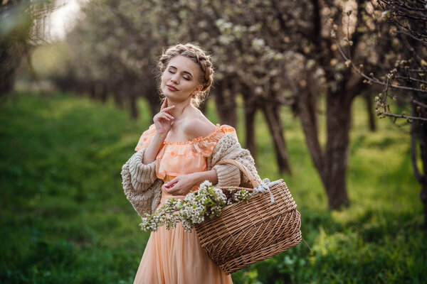 girl with blonde hair in a light dress in flowering garden. girl in a beautiful dress and knitted sweater enjoys the sunset in a pear flowering garden, with a basket of flowers