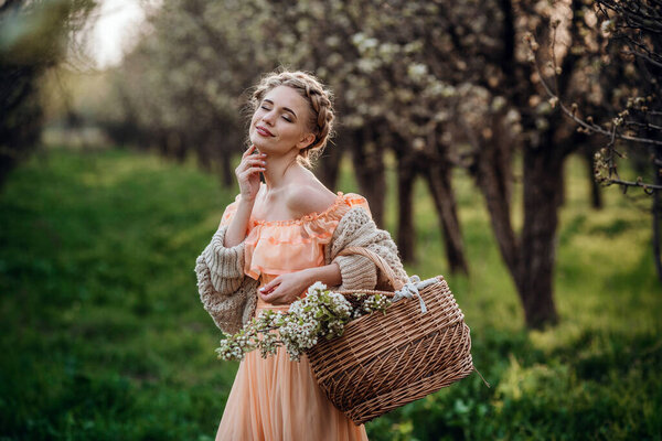 girl with blonde hair in a light dress in flowering garden. girl in a beautiful dress and knitted sweater enjoys the sunset in a pear flowering garden, with a basket of flowers
