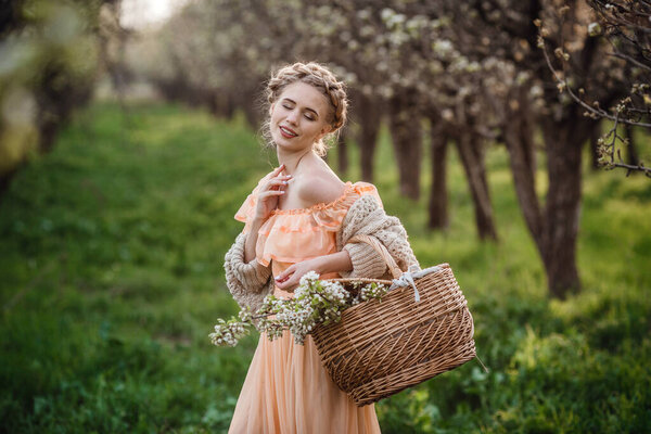 girl with blonde hair in a light dress in flowering garden. girl in a beautiful dress and knitted sweater enjoys the sunset in a pear flowering garden, with a basket of flowers