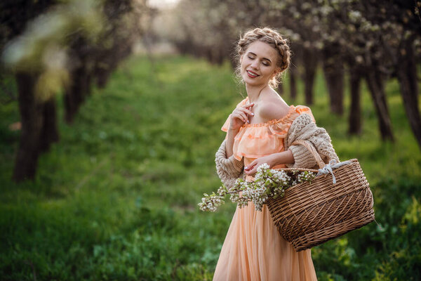 girl with blonde hair in a light dress in flowering garden. girl in a beautiful dress and knitted sweater enjoys the sunset in a pear flowering garden, with a basket of flowers