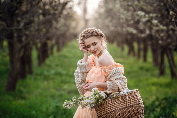 girl with blonde hair in a light dress in flowering garden. girl in a beautiful dress and knitted sweater enjoys the sunset in a pear flowering garden, with a basket of flowers