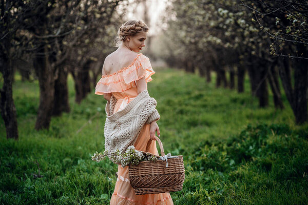 girl with blonde hair in a light dress in flowering garden. girl in a beautiful dress and knitted sweater enjoys the sunset in a pear flowering garden, with a basket of flowers