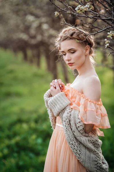 開花の庭の薄いドレスを着た金髪の女の子 女性の春のファッションの概念 美しいドレスとニットのセーターの女の子は梨の開花庭で夕日を楽しんでいます — ストック写真