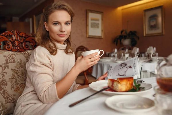 美丽的年轻女子在休息的时候坐在餐馆里看书 舒适温暖的毛衣 柔和的色调 芬芳的茶和美味的蛋糕 — 图库照片