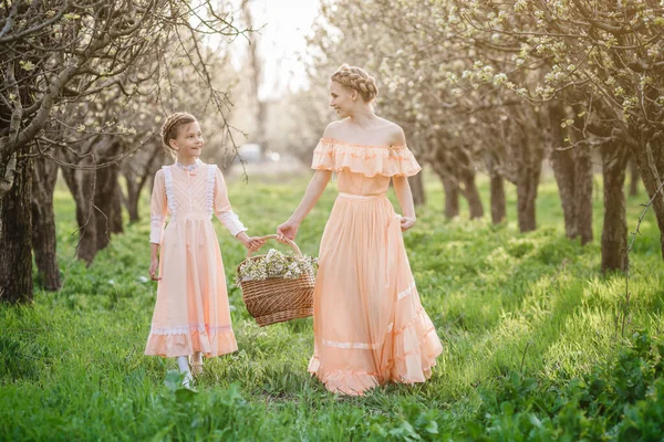 Two little sisters, younger and older, are walking in the garden, enjoying the spring in beautiful vintage dresses. The idea and concept of a friendly family and a happy childhood.