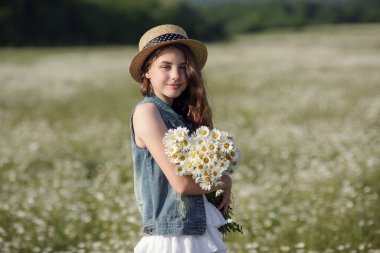 Beyaz elbiseli ve şapkalı güzel bir genç kız papatya tarlasında yürüyor. Güzel uzun ve sağlıklı saçlar. Sağlık ve alerji fikri ve konsepti, kendine dikkat etmek.