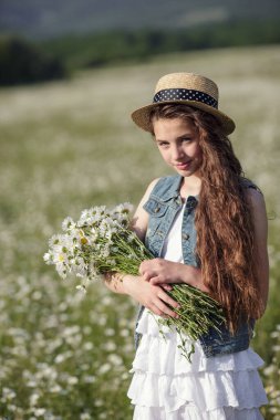 Beyaz elbiseli ve şapkalı güzel bir genç kız papatya tarlasında yürüyor. Güzel uzun ve sağlıklı saçlar. Sağlık ve alerji fikri ve konsepti, kendine dikkat etmek.
