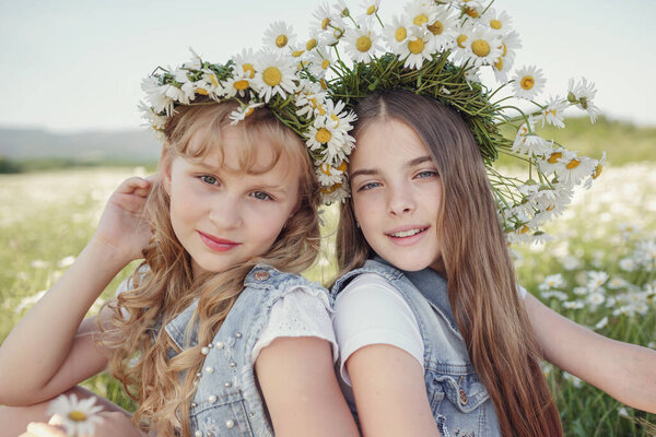two cute teen girls in denim overalls walk in a daisy field. idea and concept of health, happy childhood, friendship