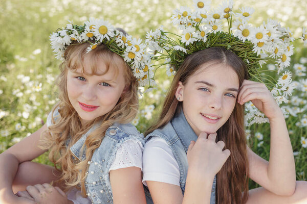 two cute teen girls in denim overalls walk in a daisy field. idea and concept of health, happy childhood, friendship