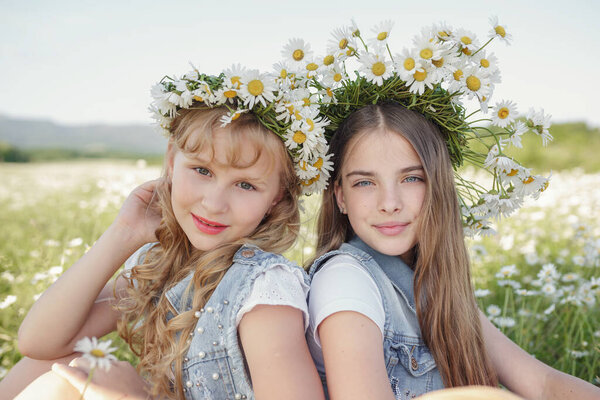 two cute teen girls in denim overalls walk in a daisy field. idea and concept of health, happy childhood, friendship
