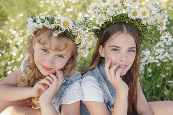 two cute teen girls in denim overalls walk in a daisy field. idea and concept of health, happy childhood, friendship
