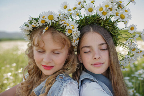 two cute teen girls in denim overalls walk in a daisy field. idea and concept of health, happy childhood, friendship