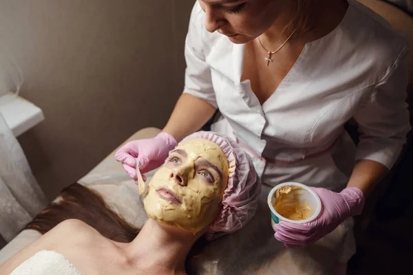 Cosmetology specialist applying gold facial mask using brush, making skin hydrated and face glowing and skin. Attractive brunette relaxing and enjoying spa procedures. Beauty and relaxation concept.