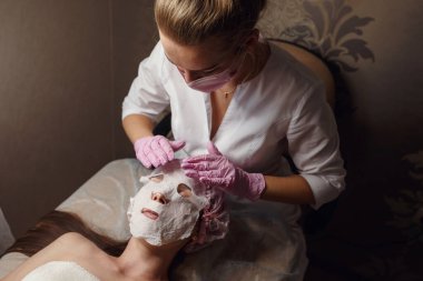 Güzellik tedavisi konsepti. Yüzüne beyaz nemlendirici kumaş maskeli güzel bir kadın. Spa, cilt bakımı, cilt temizleme ve nemlendirici prosedürler. Sağlık ve rahatlama..