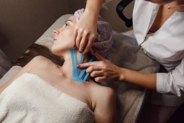 Kadın kozmetikçi, evdeki masada yatan hastanın yüzünü bantlıyor. Fizyoterapi ve kozmetoloji prosedürü. Cerrahi olmayan cilt yenileme metodu. Yüz bakımı.