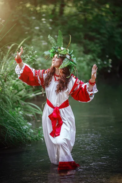 beautiful woman in wreath of wild flowers of flowers in sunny day. Pagan Slavic adult woman in a white dress with red national embroidery conduct ceremony on Midsummer. Earth Day or Ivana Kupala