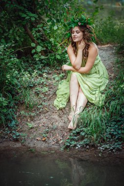 Yeşil elbiseli güzel bir kadın ve güneşli bir ormanda çelenk. Yaz ortası ya da Dünya Günü 'nde seremoni. Parfüm fikri ve konsepti, peri masalları ve mutluluk