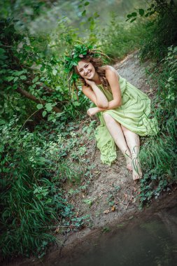 Yeşil elbiseli güzel bir kadın ve güneşli bir ormanda çelenk. Yaz ortası ya da Dünya Günü 'nde seremoni. Parfüm fikri ve konsepti, peri masalları ve mutluluk