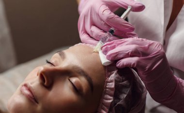 Kadın güzellik kliniğinde gençleştirme prosedürü uyguluyor. Klinikte sakince yatıyor. Pembe eldivenli uzman güzellik uzmanı hyaluronik asitle kadın kırışıklıklarını dolduruyor. Güzellik aşısı.