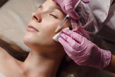 Kadın güzellik kliniğinde gençleştirme prosedürü uyguluyor. Klinikte sakince yatıyor. Pembe eldivenli uzman güzellik uzmanı hyaluronik asitle kadın kırışıklıklarını dolduruyor. Güzellik aşısı.